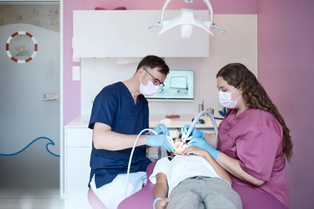 Zahnarzt und Assistentin behandeln gemeinsam die Zähne eines liegenden Jungen im rosa Behandlungszimmer