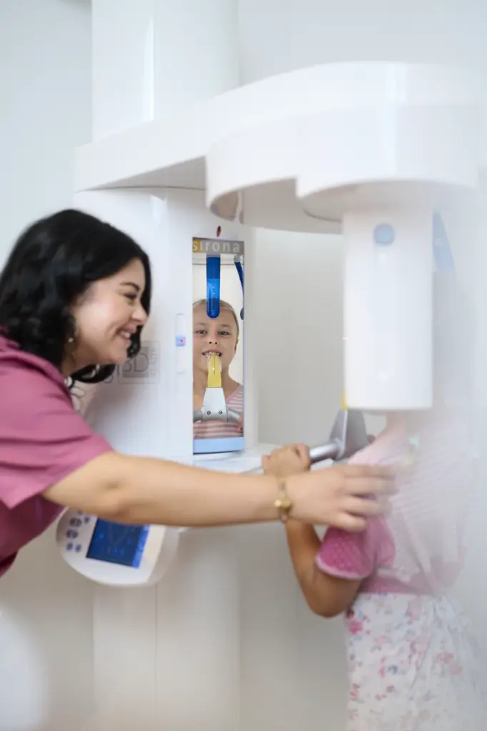 Mitarbeiterin positioniert lächelnd ein kleines Mädchen für eine digitale Röntgenaufnahme im 3D-Röntgengerät