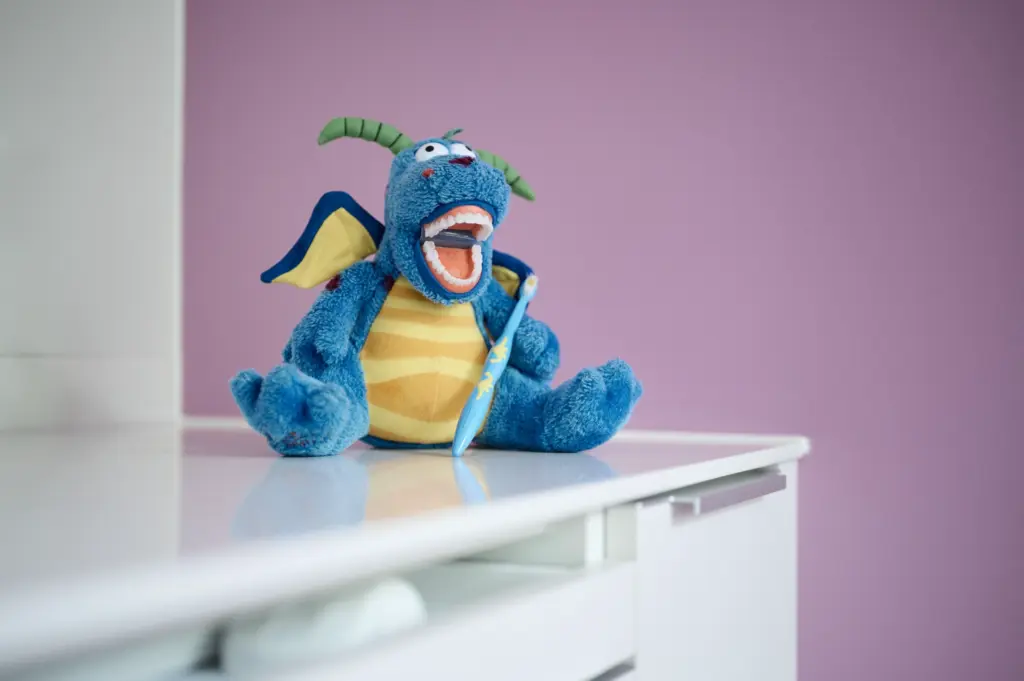 Blauer Plüsch-Drache mit weit geöffnetem Mund und Zahnbürste sitzt auf einer weißen Ablage vor rosa Wand
